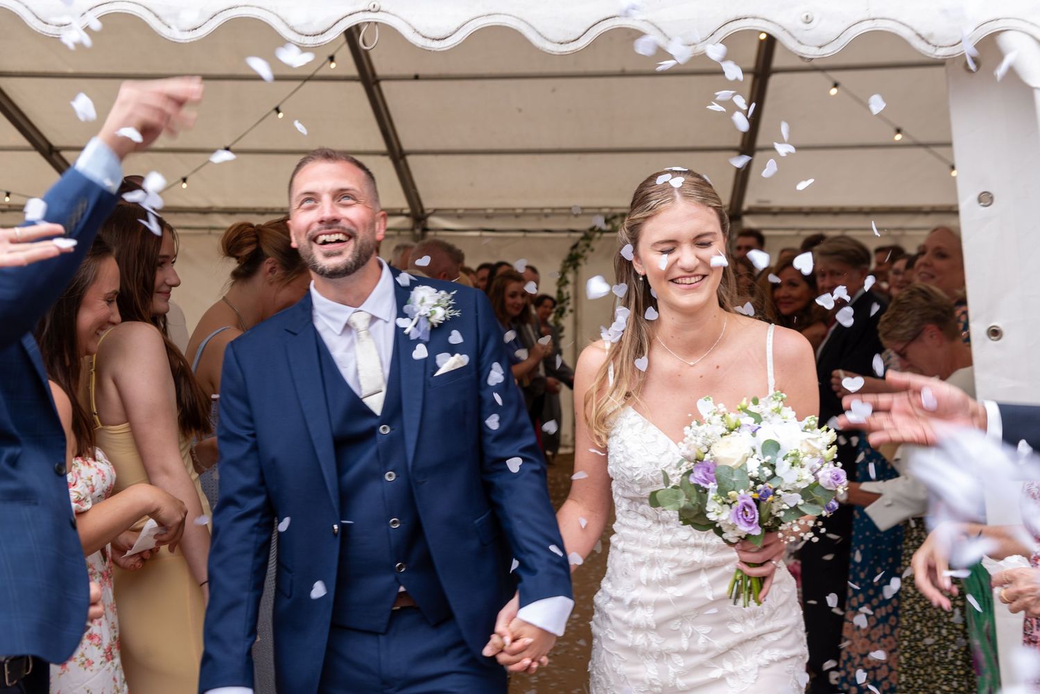 Bride and groom laughing as confetti falls during wedding exit under marquee tent.