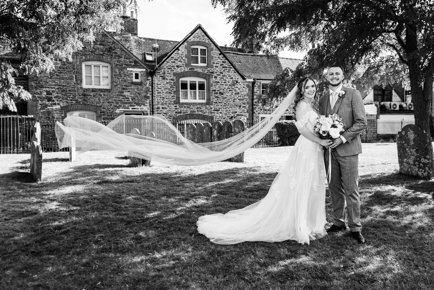 Black and white wedding photo of bride and groom posing outdoors, veil flowing in wind, stone building backdrop.