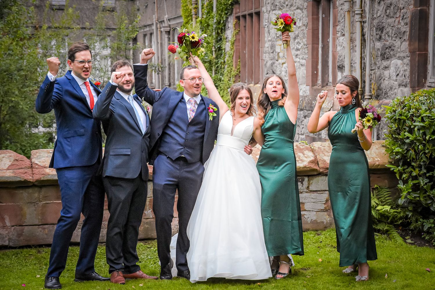 Joyful wedding party celebrating outdoors, bride in white gown with bridesmaids in green dresses and groomsmen in suits.