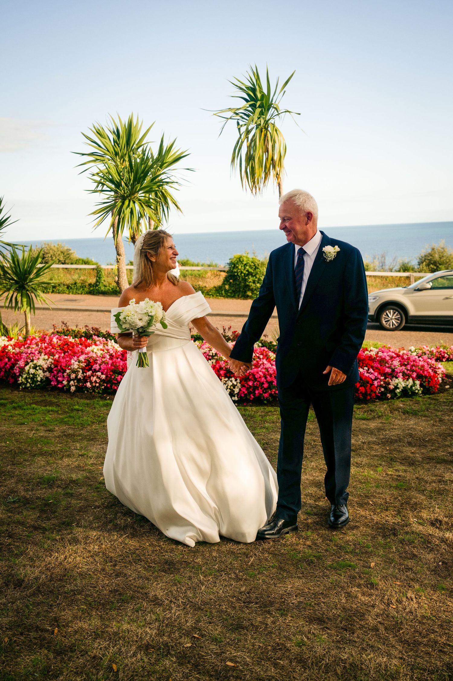 A bride and groom hold hands and smile while walking on grass, with colorful flowers and palm trees in the background.