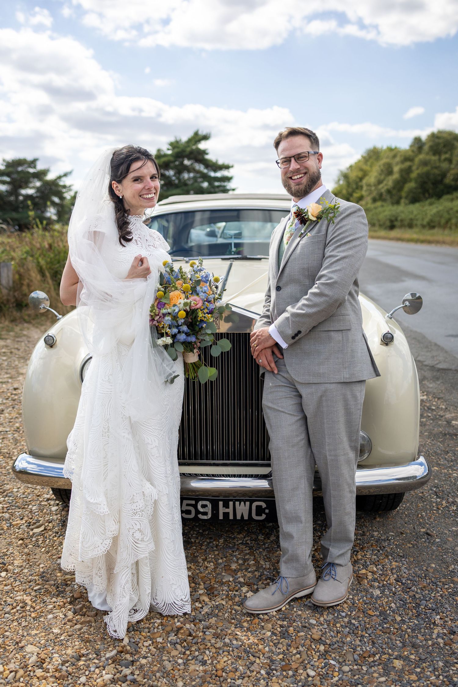 Bride and groom smiling beside a vintage cream Rolls-Royce, bride holding colorful bouquet, groom in grey suit.