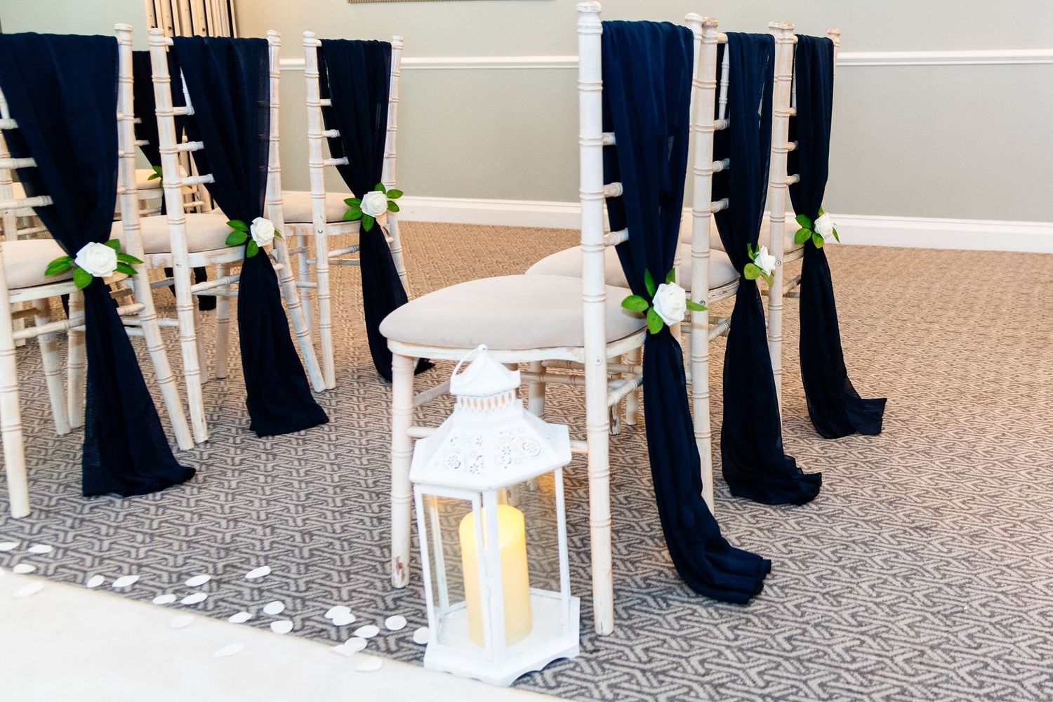 Elegant wedding ceremony chairs adorned with navy blue sashes and white roses, flanking a white lantern centerpiece.