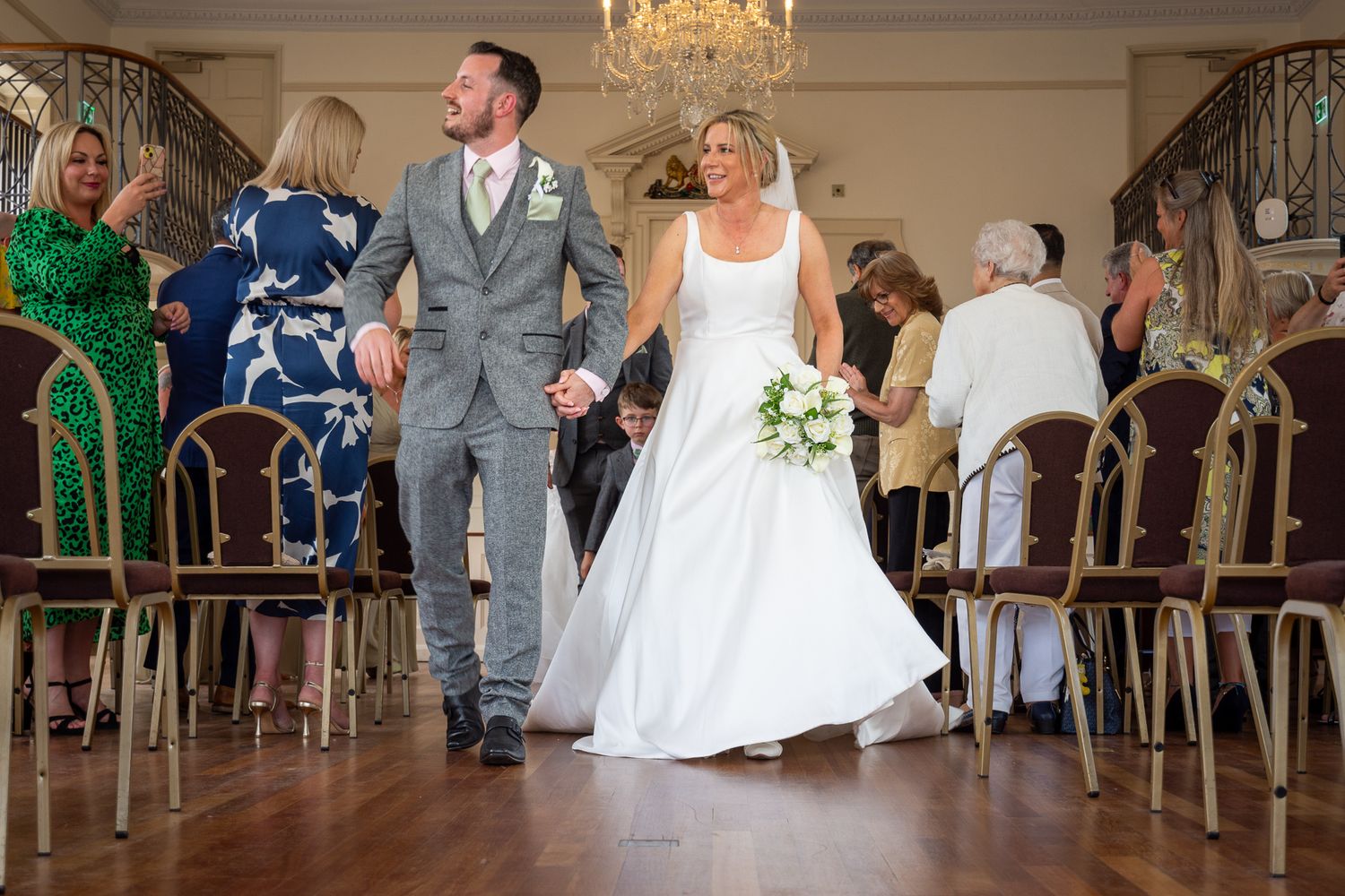 Bride and groom walk hand-in-hand down the aisle after wedding ceremony in elegant venue with guests applauding.
