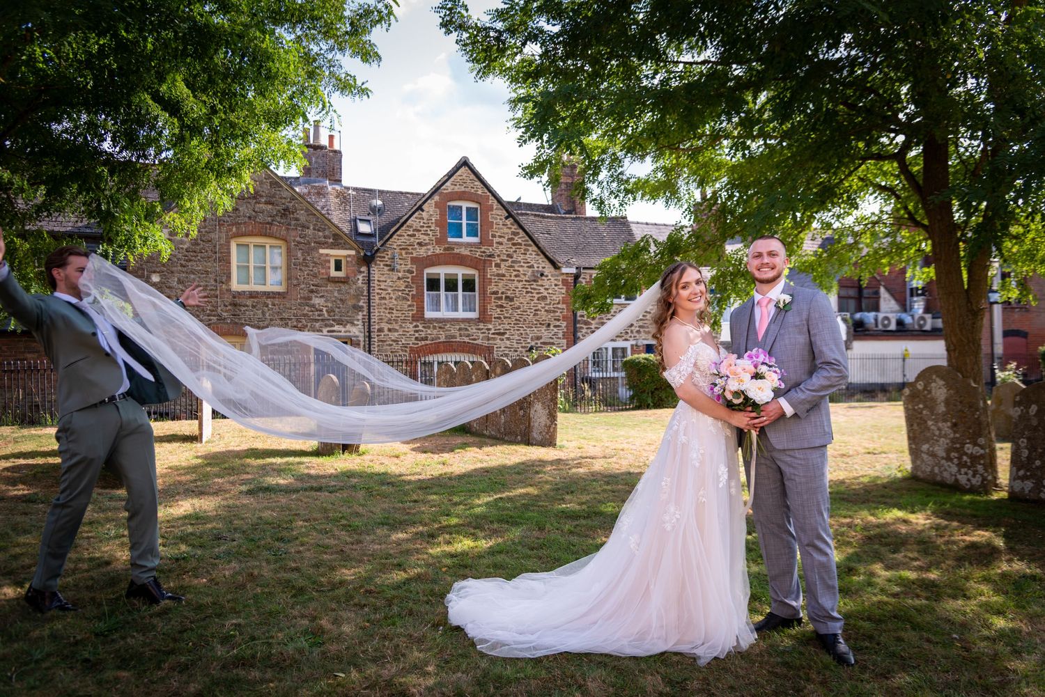 Bride and groom pose outside stone building while groomsman dramatically extends flowing wedding veil.