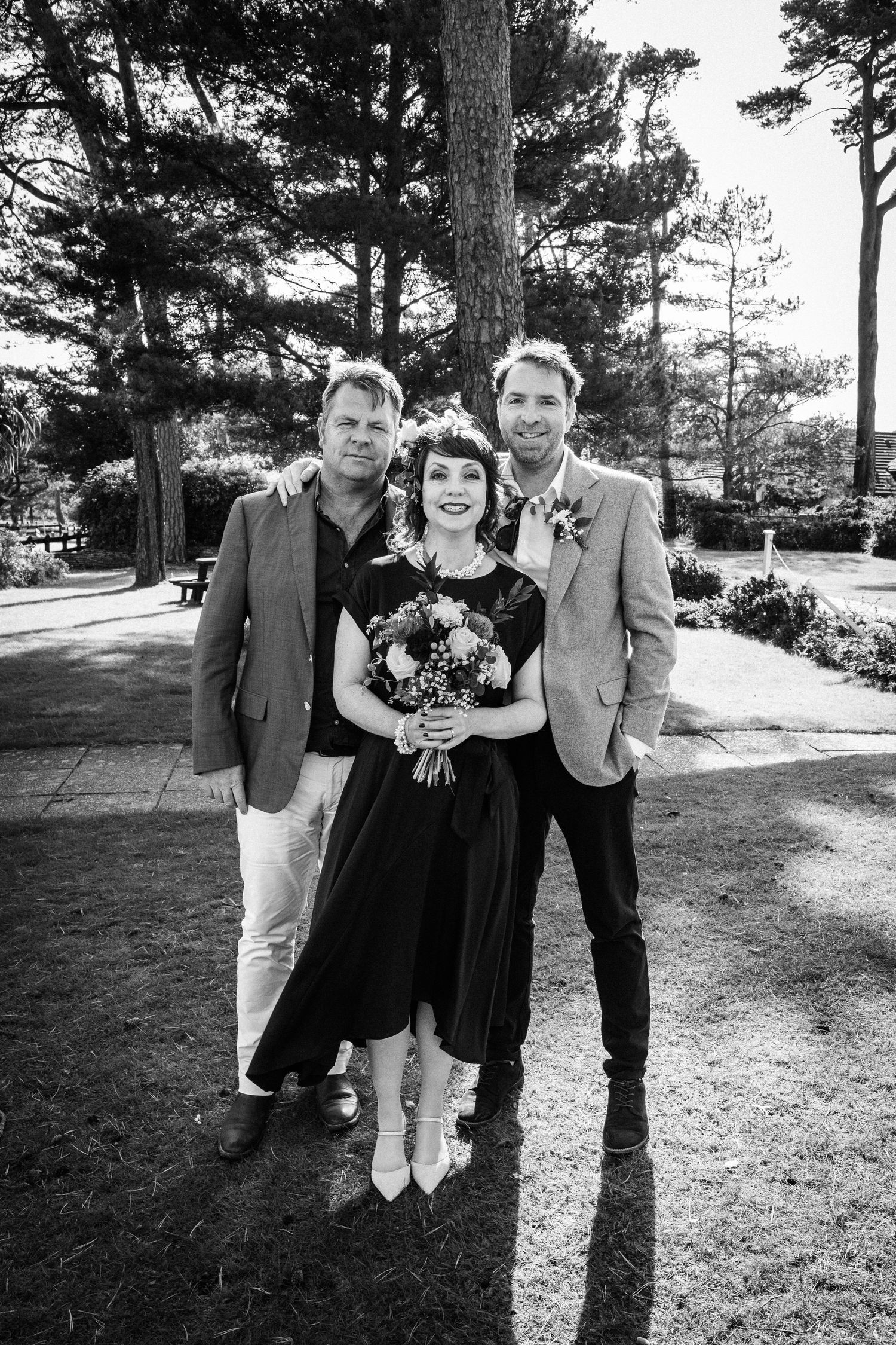 Black and white photo of a bride in black dress holding bouquet posing with two men in blazers outdoors.