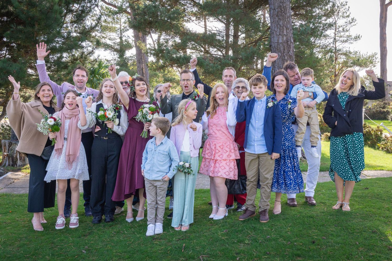 Large wedding party group photo outdoors on green lawn with trees, guests cheering and celebrating together.