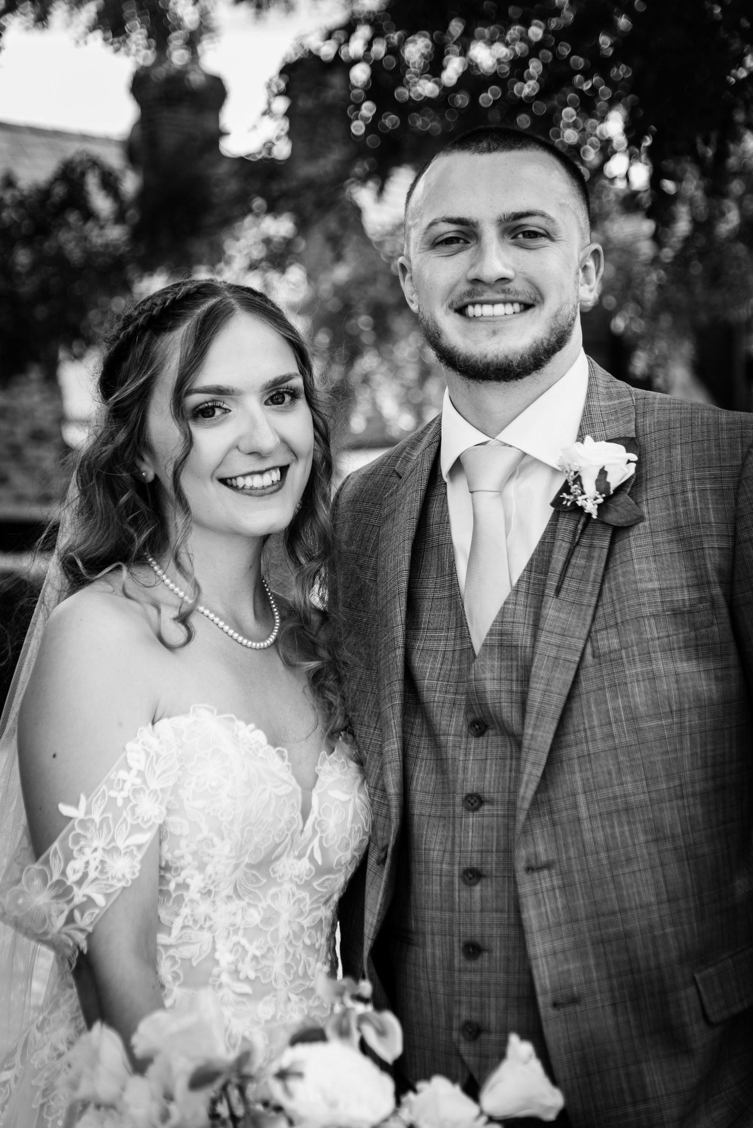 Black and white wedding portrait of smiling bride in lace gown with bouquet and groom in plaid suit with boutonniere.
