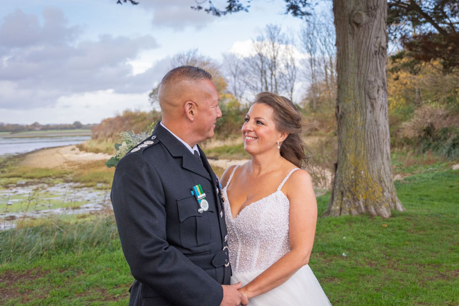 Bride and groom in military uniform holding hands outdoors near a lake and trees on their wedding day.