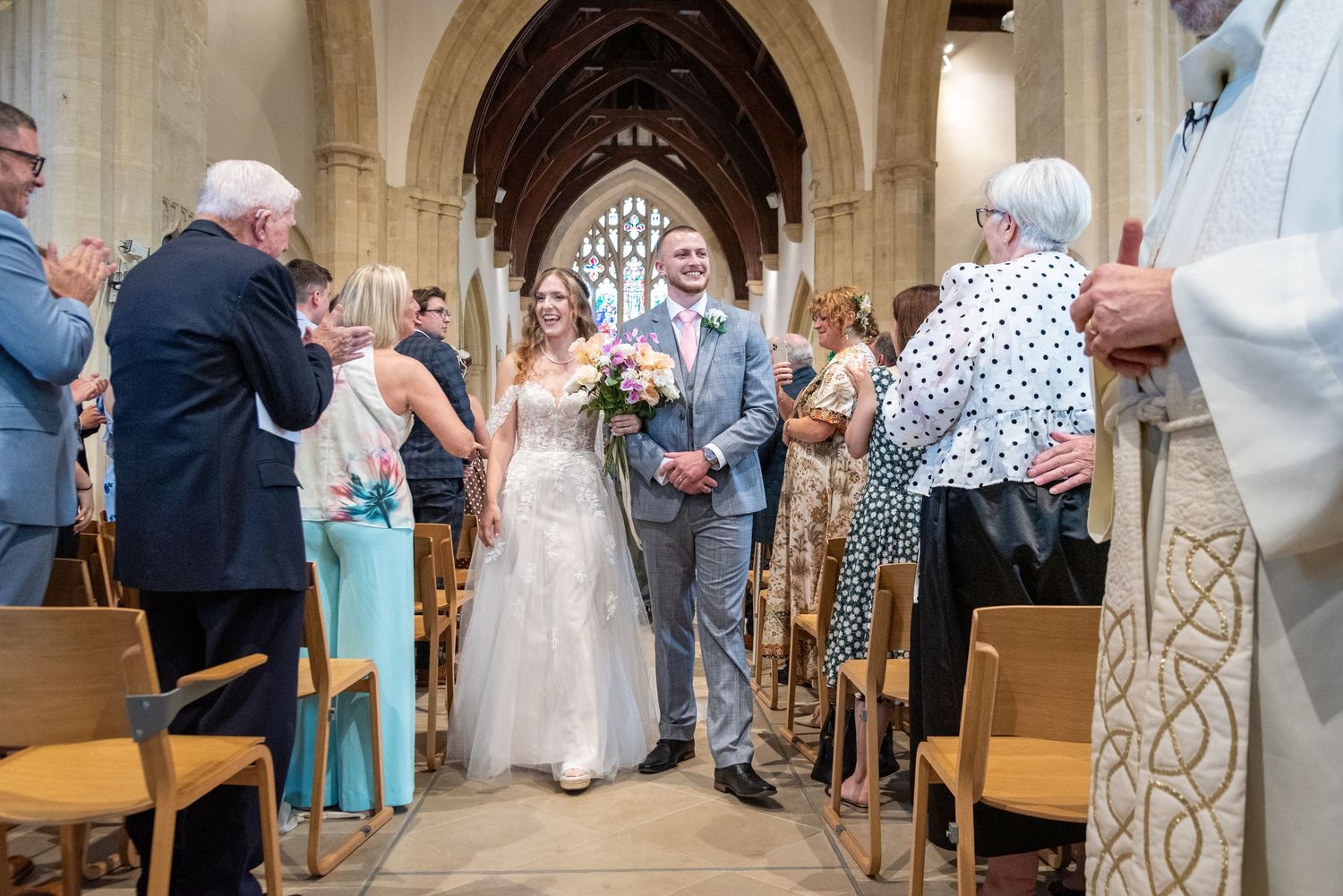 Bride and groom walk down church aisle after wedding ceremony, greeted by smiling guests in pews.