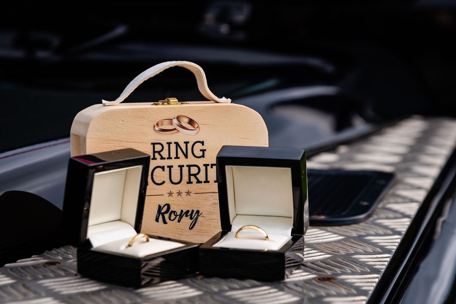 Wooden ring bearer box engraved with Ring Curi and Roxy beside a black velvet ring box with gold wedding bands.
