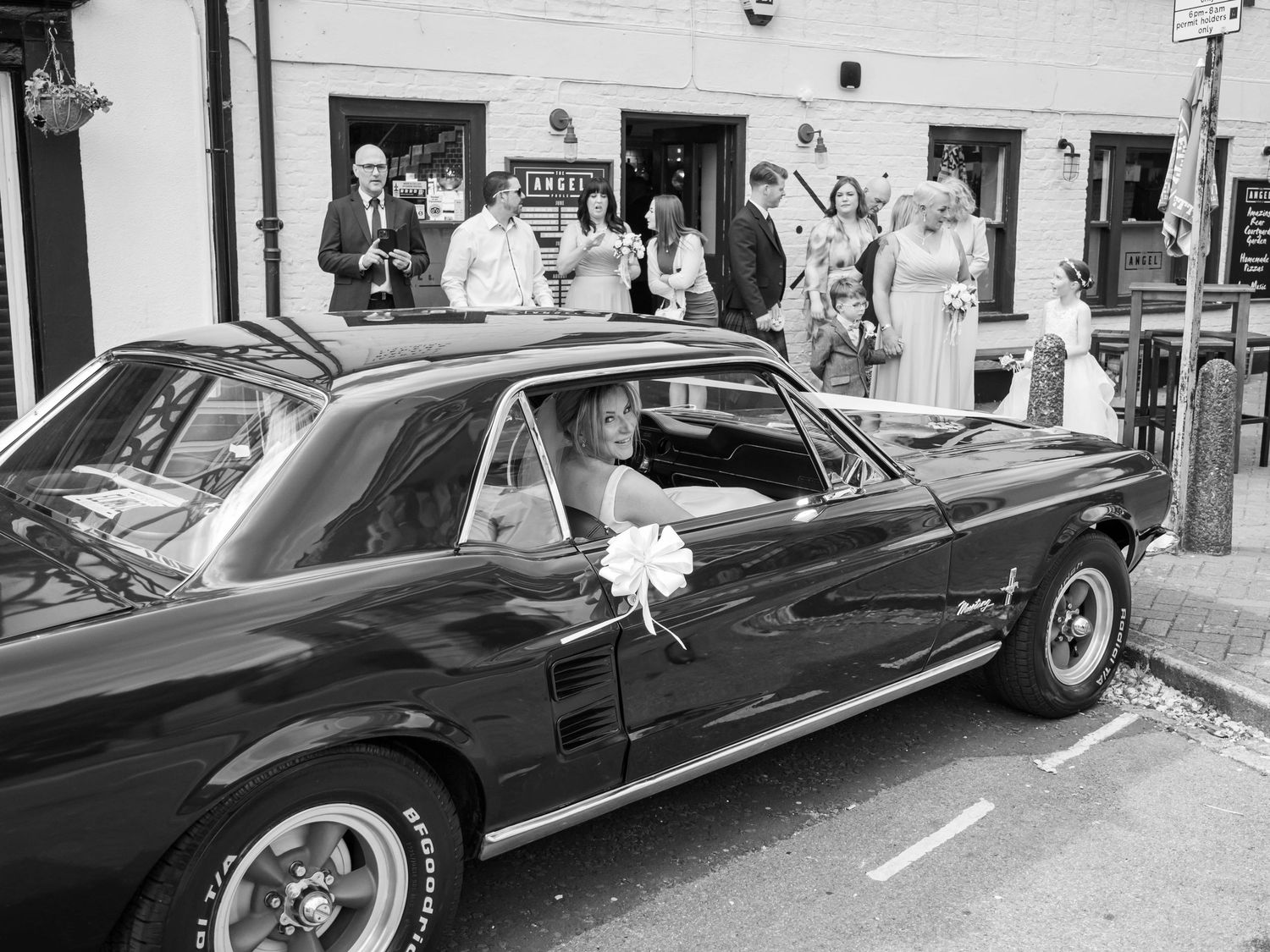 Black and white photo of a bride in a classic Ford Mustang wedding car with guests outside a venue.