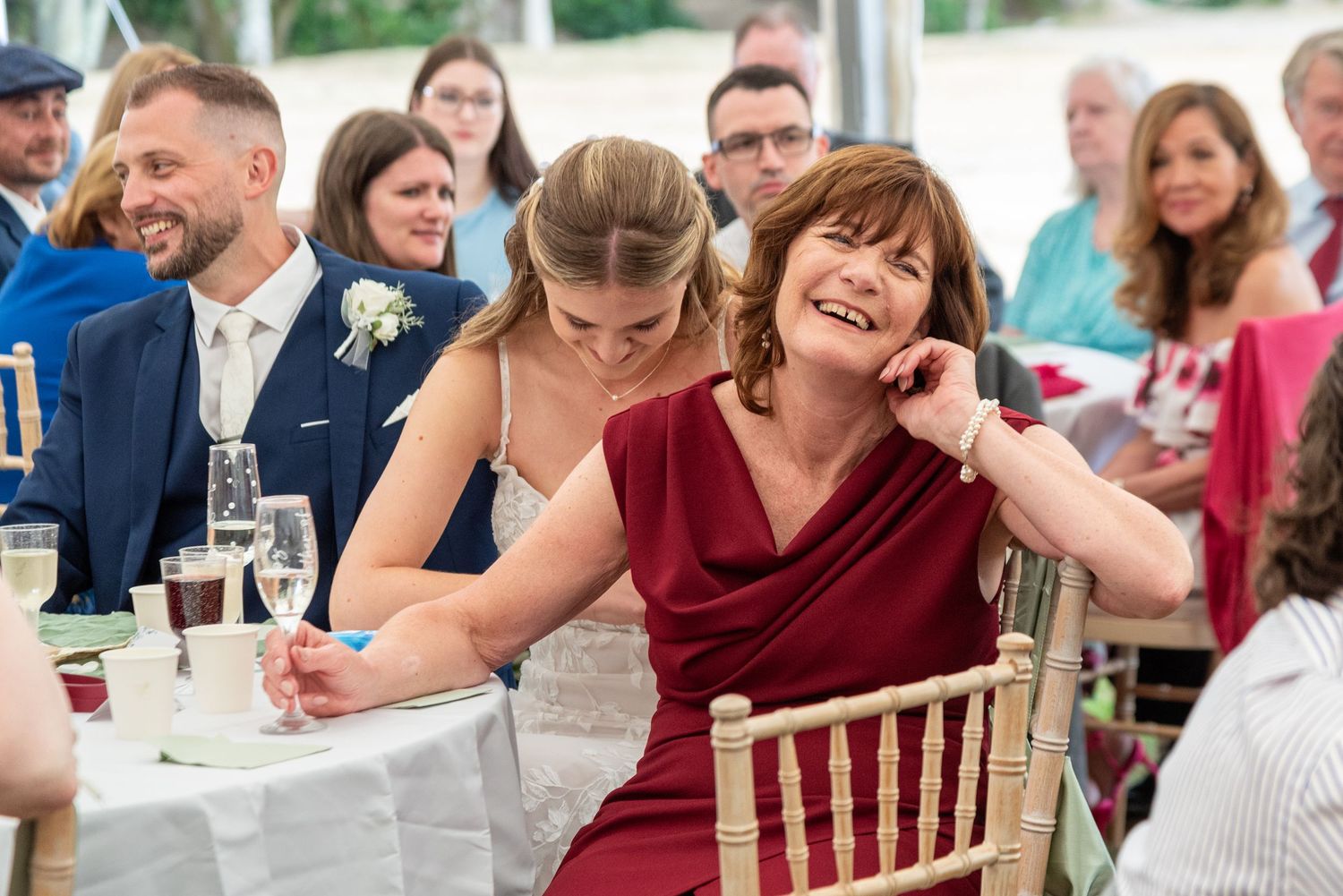 Guests laughing and enjoying wedding reception speeches at an elegant outdoor marquee celebration.