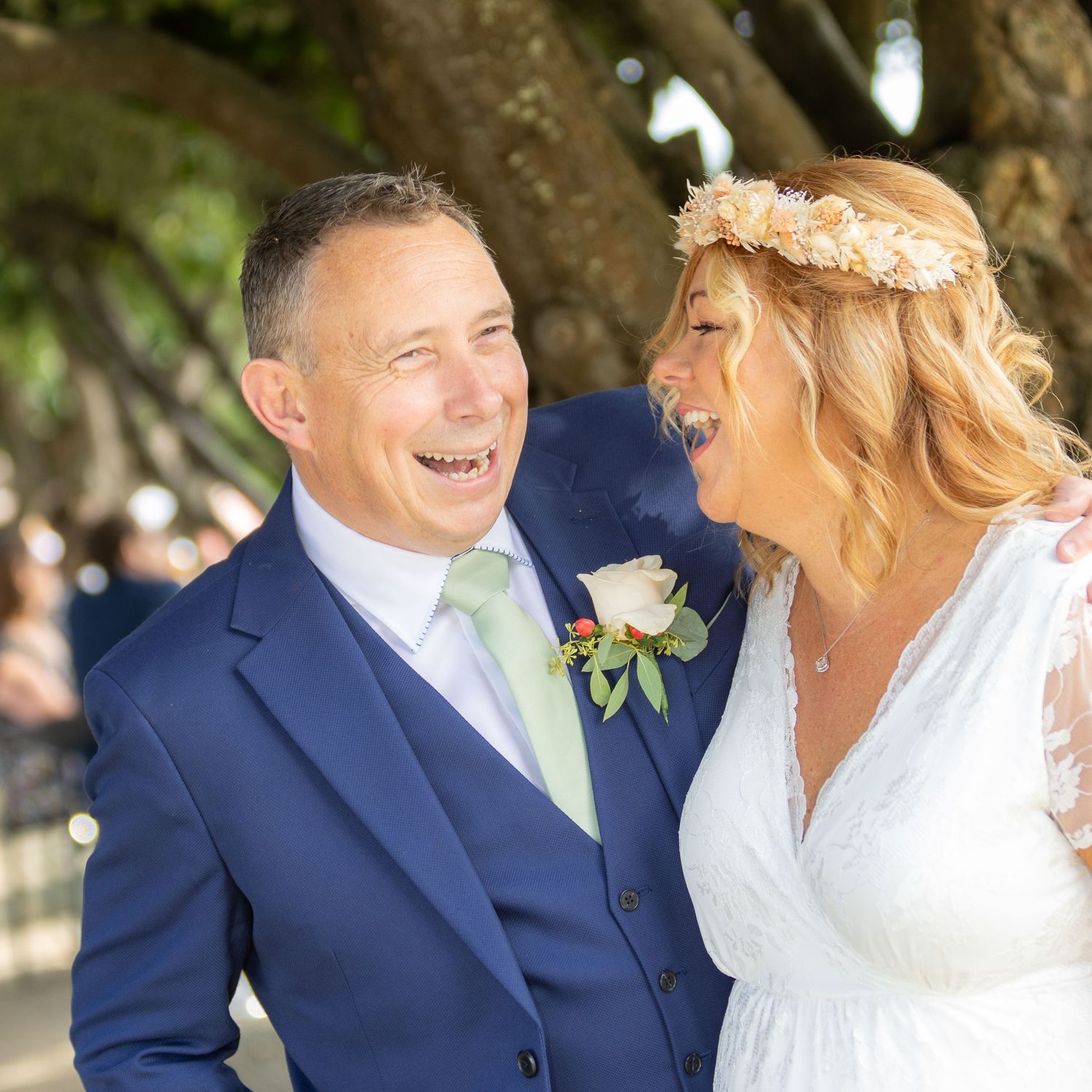 A joyful couple laughing together on their wedding day, bride in white dress with floral crown, groom in navy blue suit.