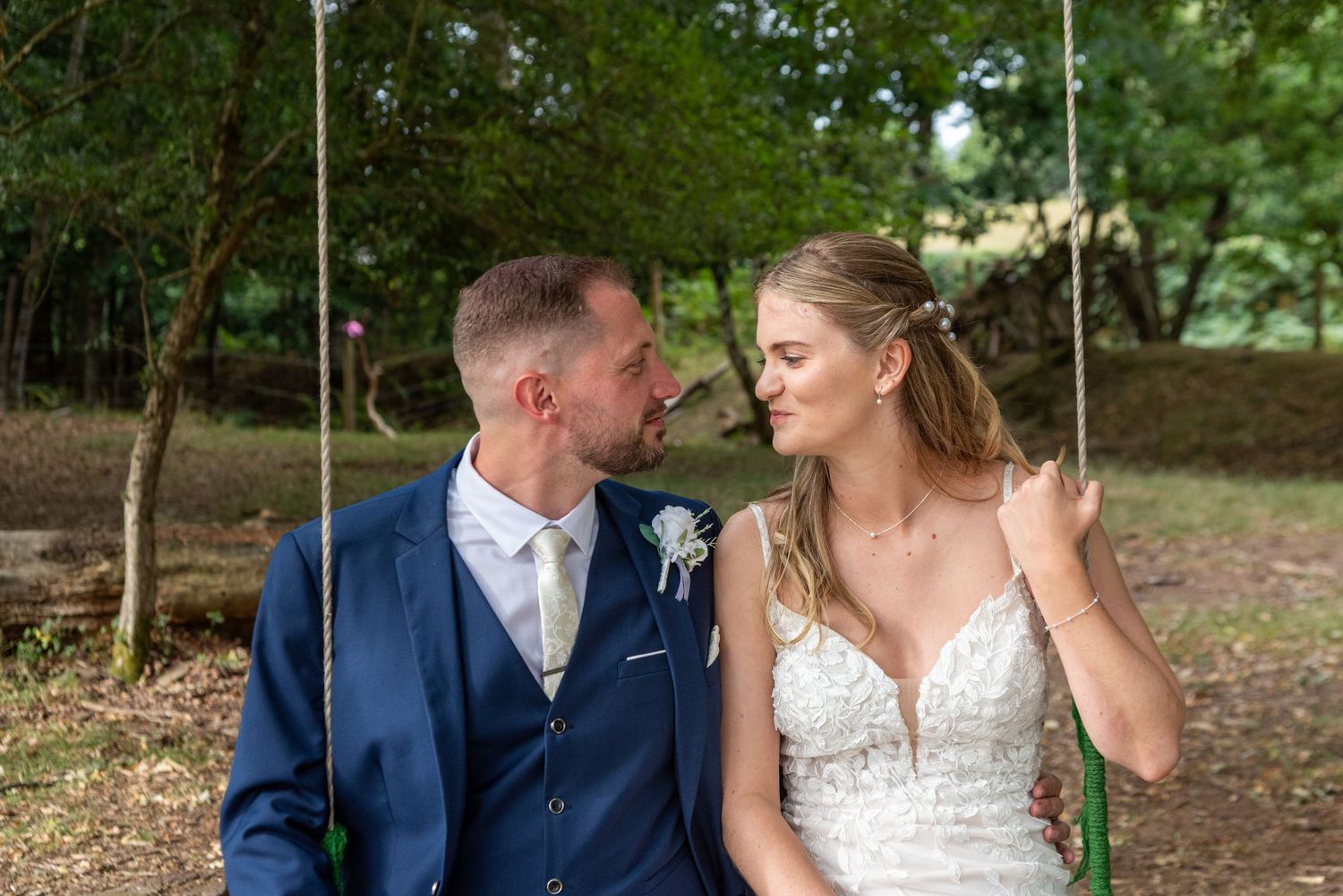 A bride and groom sit together on a swing, smiling at each other in a lush outdoor woodland setting.