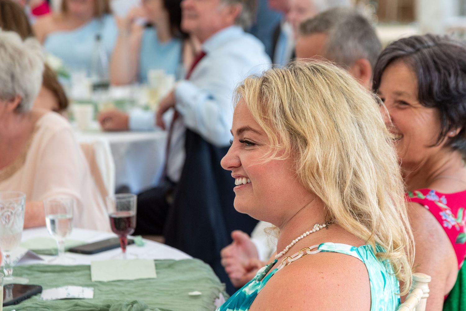 Smiling blonde woman in floral dress enjoys wedding reception with guests at decorated tables.