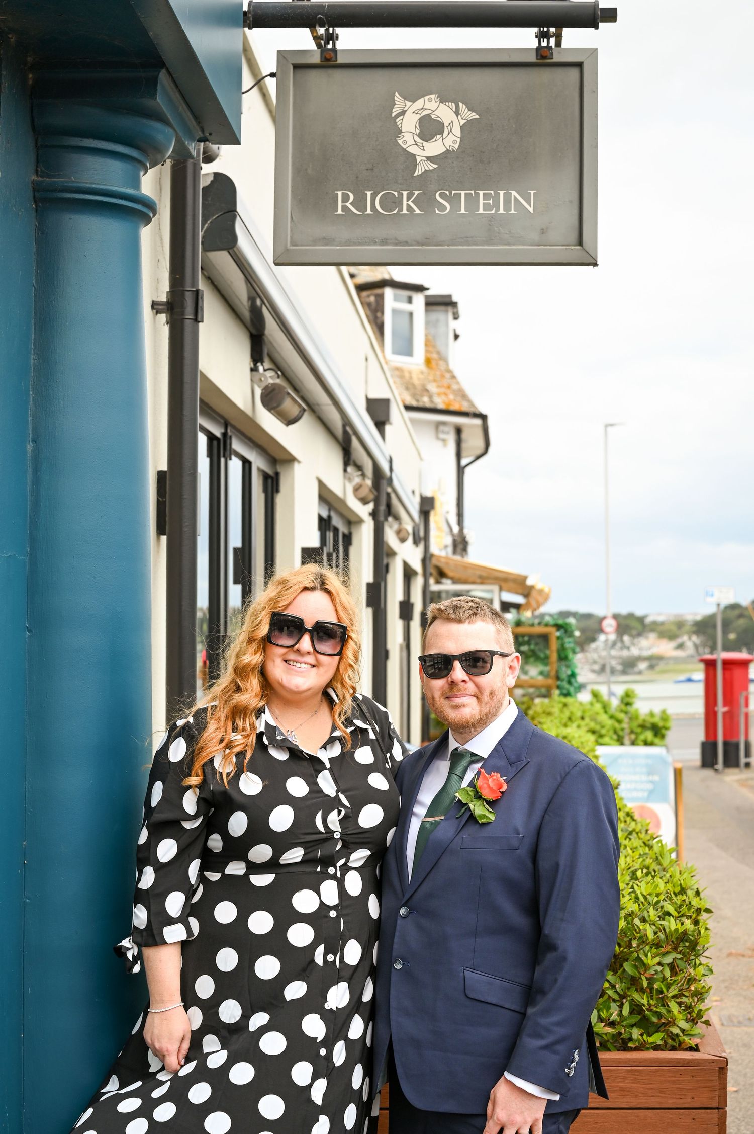 Couple posing outside Rick Stein restaurant, woman in polka dot dress and man in navy suit with boutonniere.