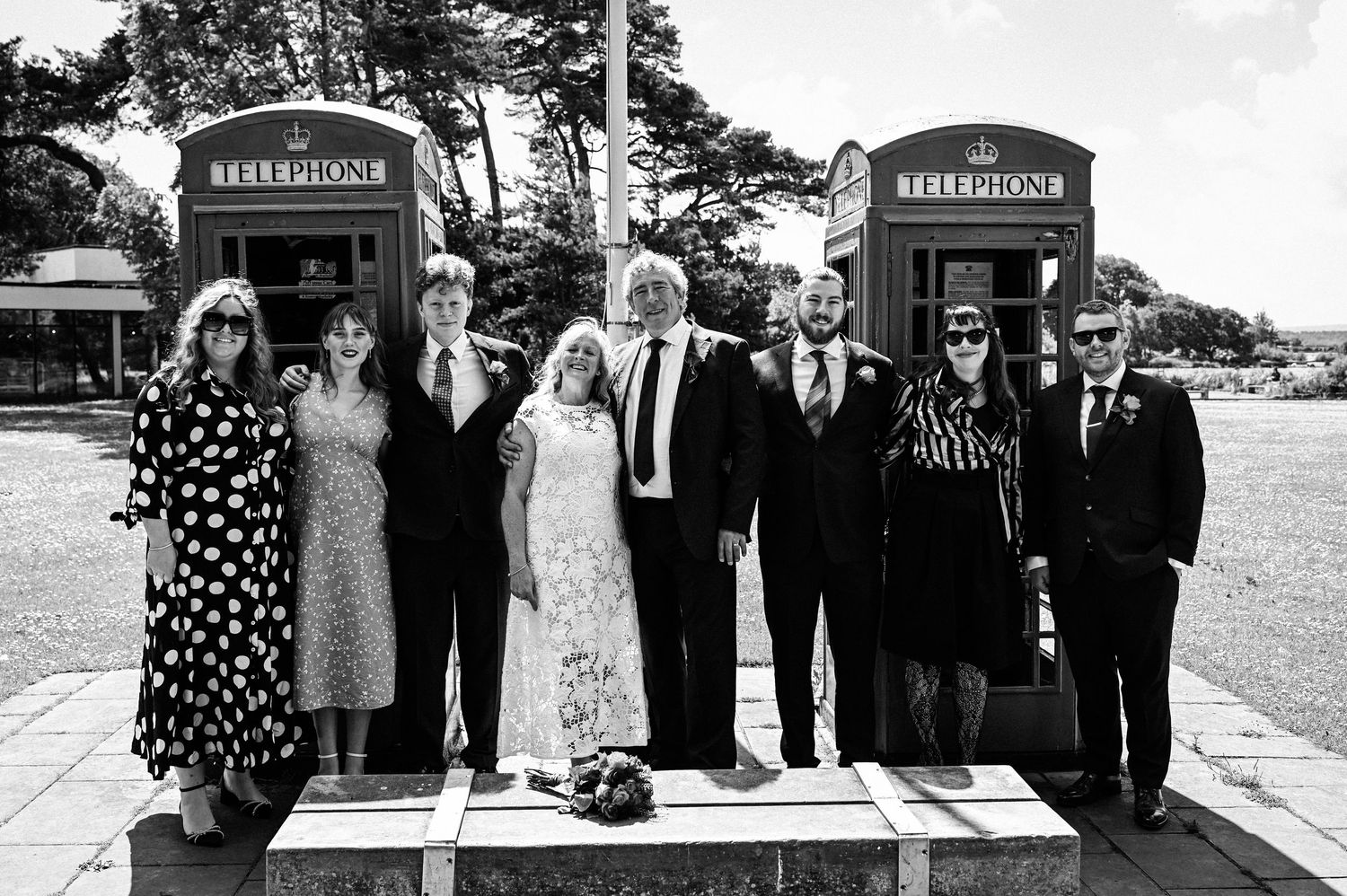 Black and white photo of a wedding party posing in front of red British telephone boxes outdoors.