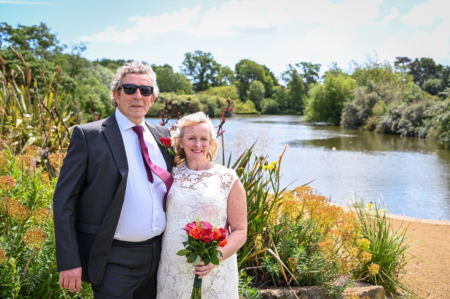 Bride and groom posing by a scenic lake surrounded by colorful flowers on their wedding day.