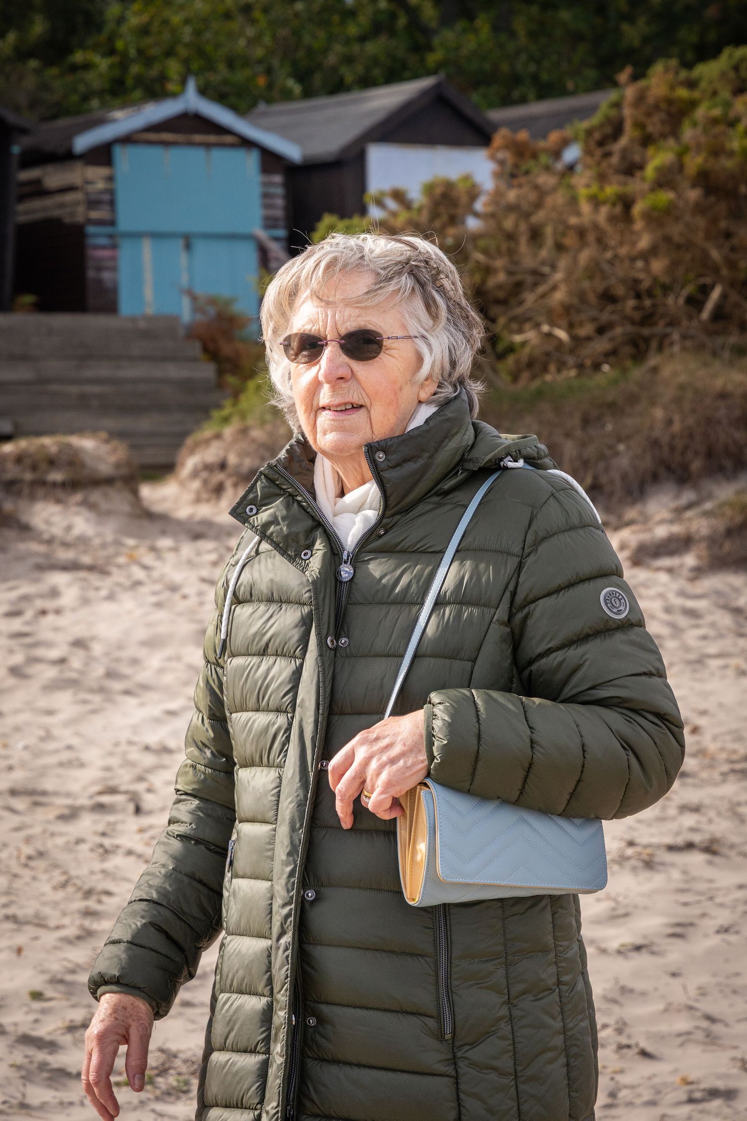 Elderly woman in green puffer jacket and sunglasses walking on sandy beach with colorful beach huts in background.