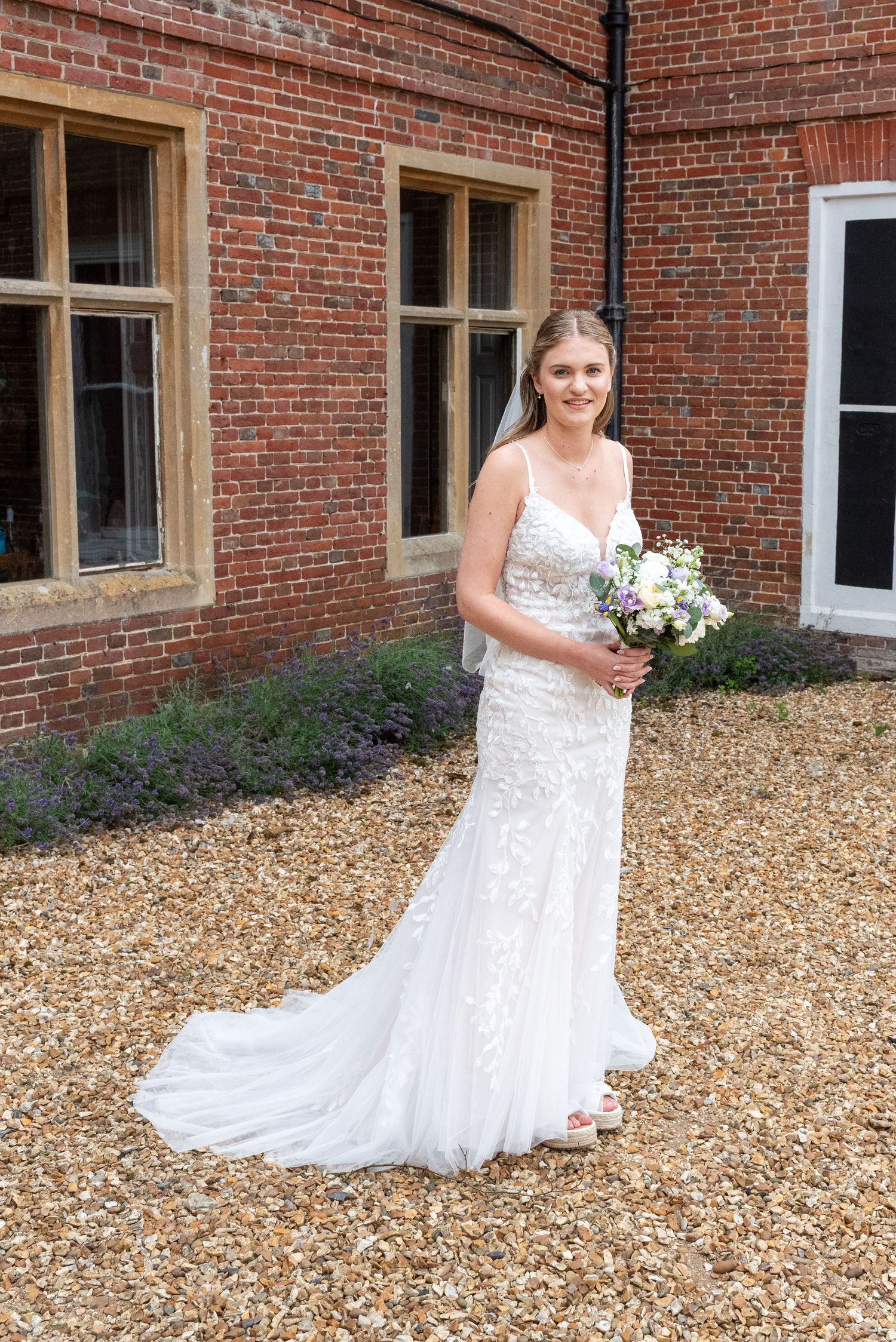 Bride in white lace wedding gown holding bouquet stands on gravel outside a red brick venue.