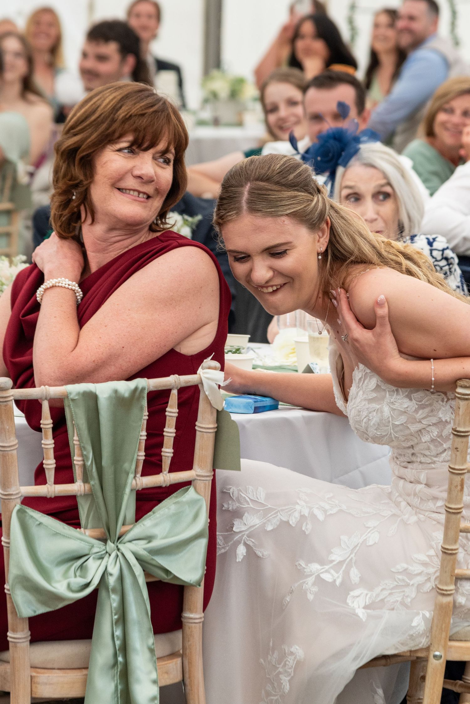 Bride in lace gown leaning affectionately toward older woman in burgundy dress at reception.