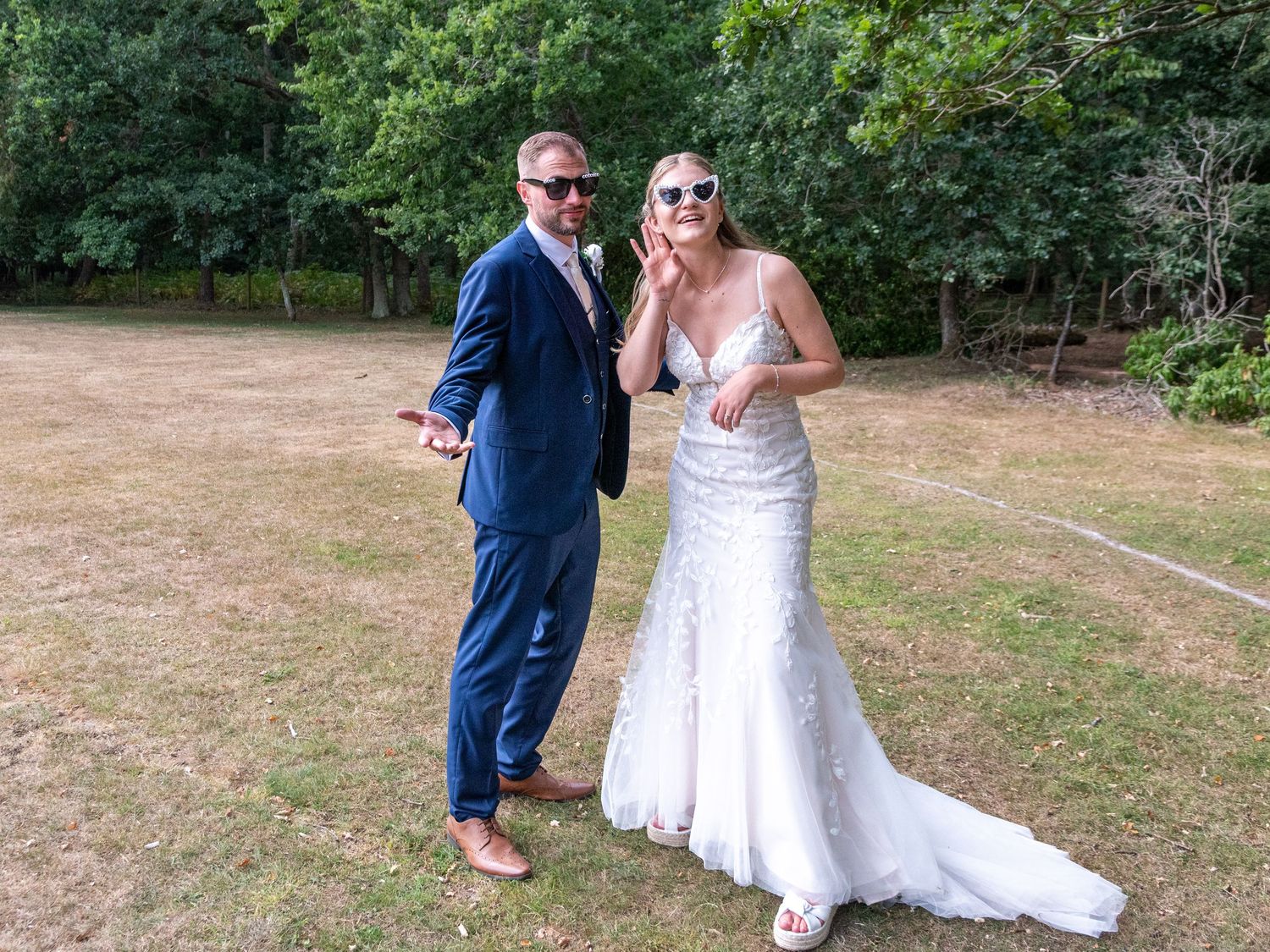 Bride in fitted lace gown and groom in navy suit pose playfully outdoors on a sunny wedding day.
