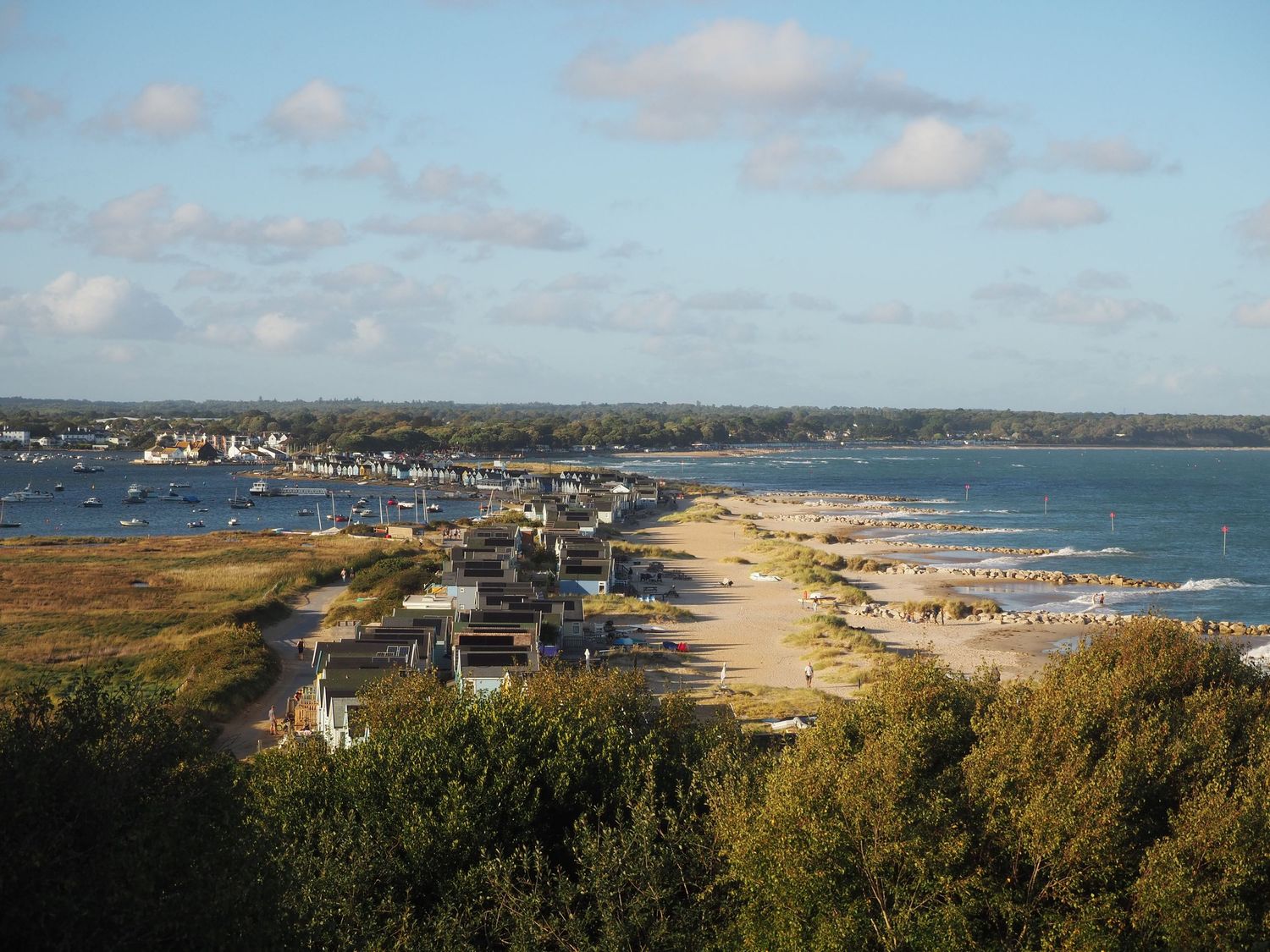 Aerial view of a coastal town with sandy beach, calm bay waters, and lush green trees under a partly cloudy sky.