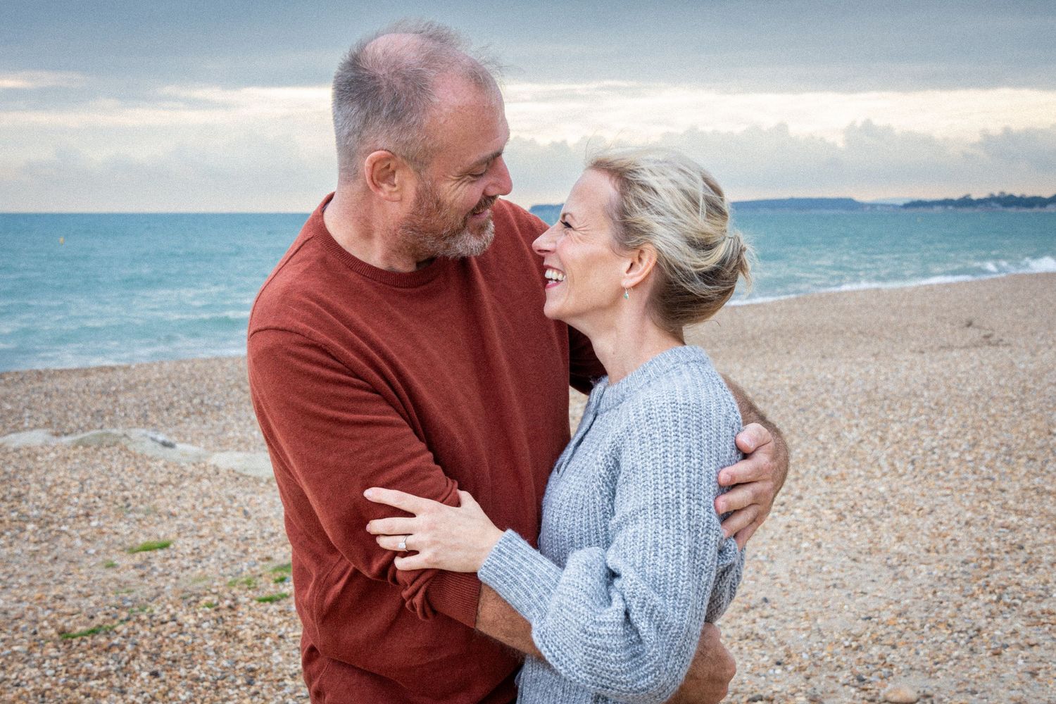 Middle-aged couple embracing and smiling at each other on a pebble beach with calm ocean waves in the background.