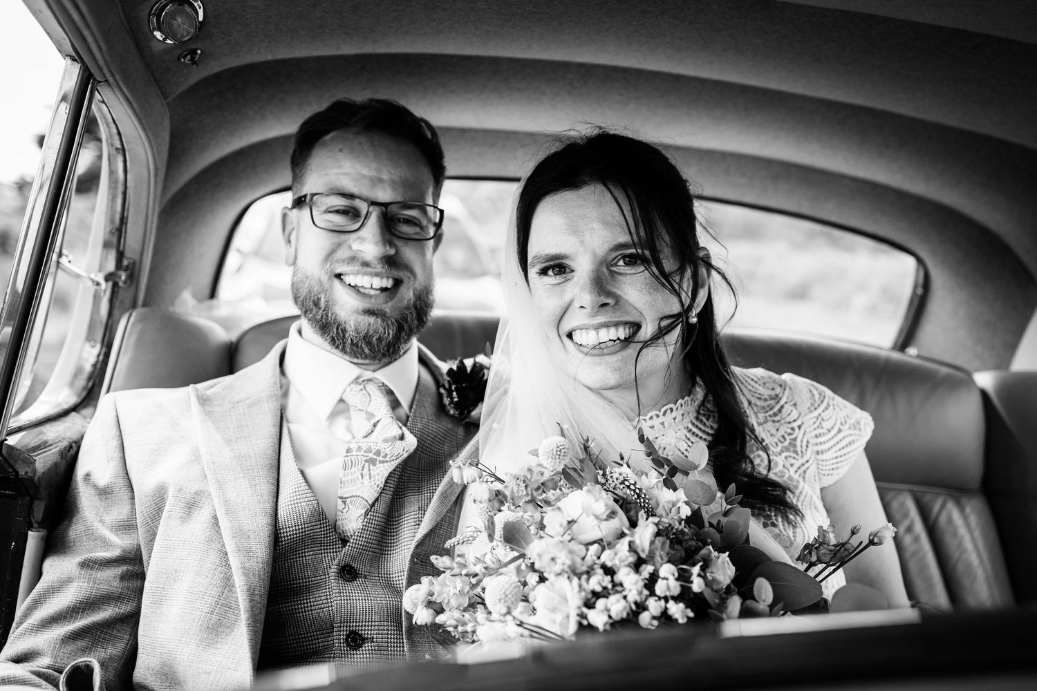 Smiling bride and groom sit together in vintage car backseat, bride holding bouquet, in black and white wedding photo.