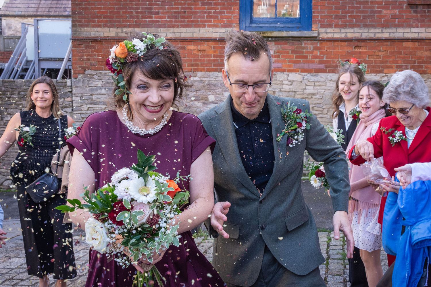 Bride and groom walk through confetti shower after wedding ceremony, bride wearing floral crown and holding bouquet.