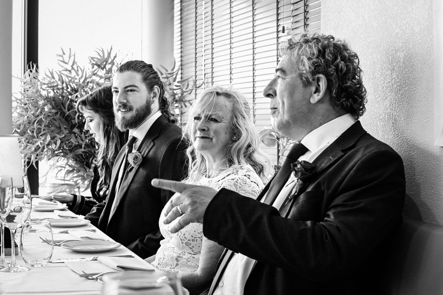 Black and white photo of three wedding guests seated at a reception table, smiling and gesturing during speeches.