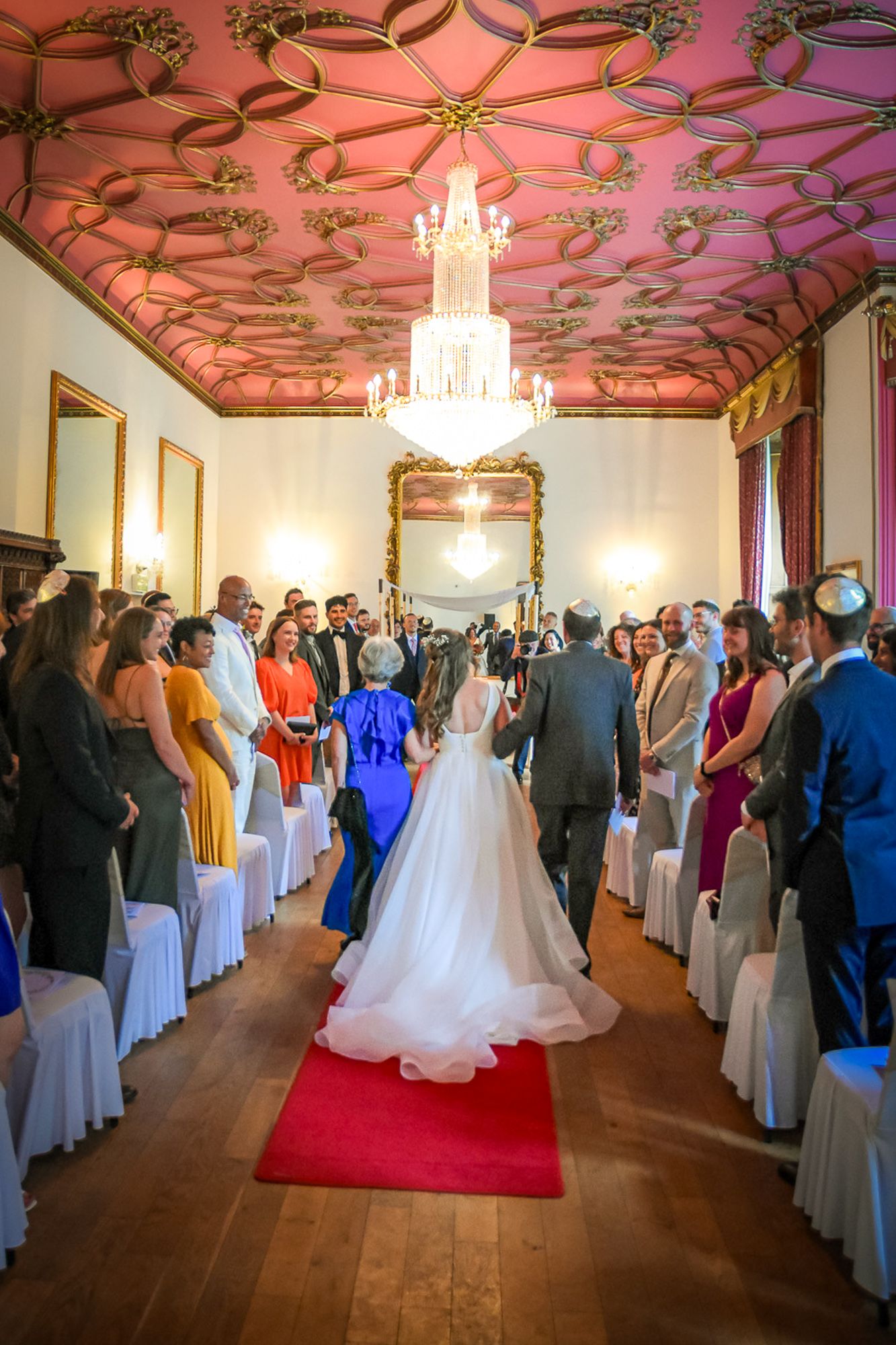 Elegant wedding ceremony in ornate hall with red ceiling, chandelier, guests lining red carpet aisle as bride walks forward.