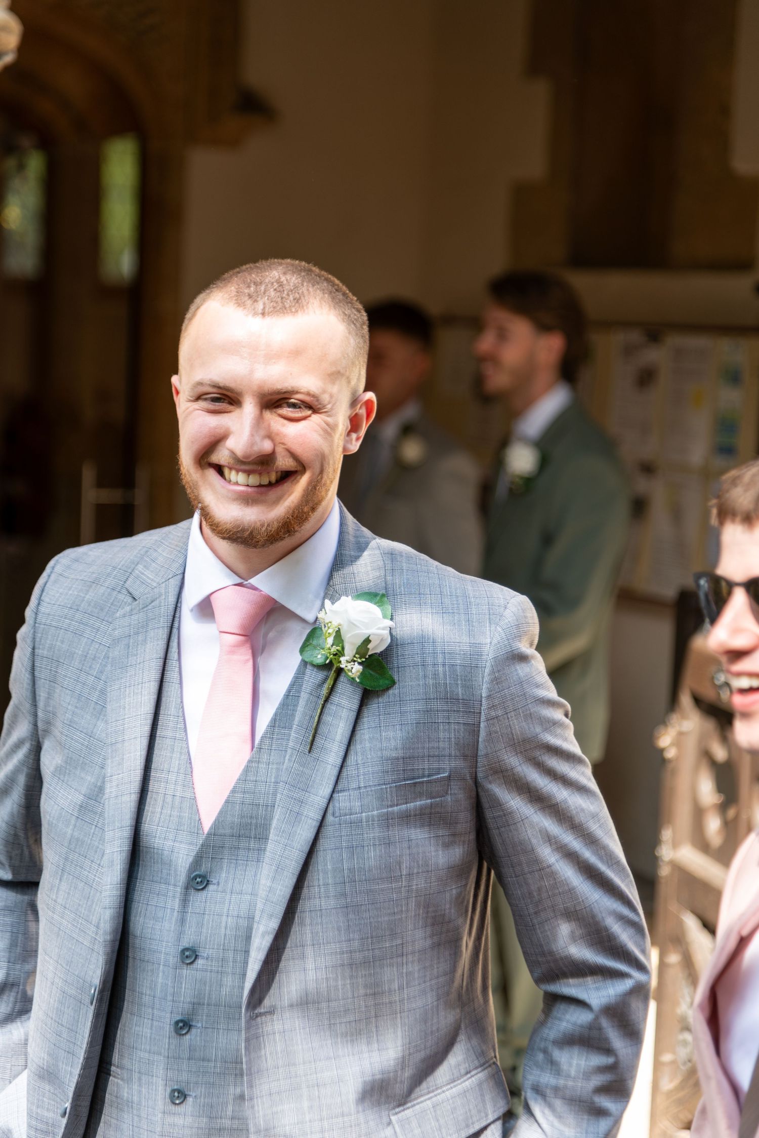 Smiling groom in grey suit with pink tie and white boutonniere at outdoor wedding ceremony.
