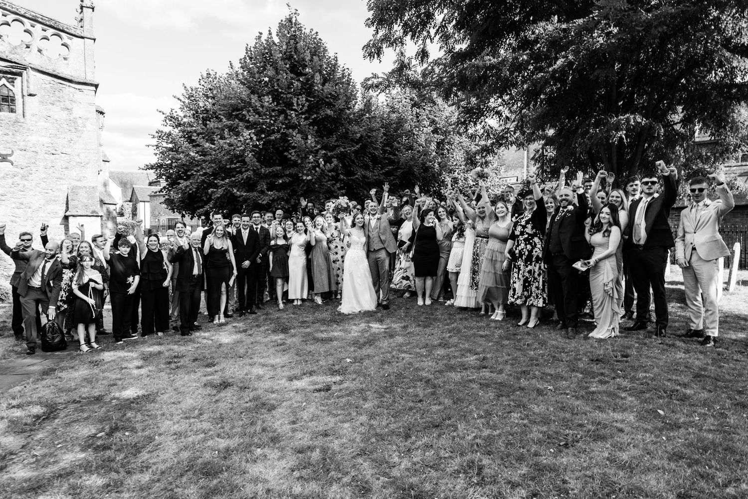 Black and white wedding group photo with large crowd of guests celebrating outdoors near a church.