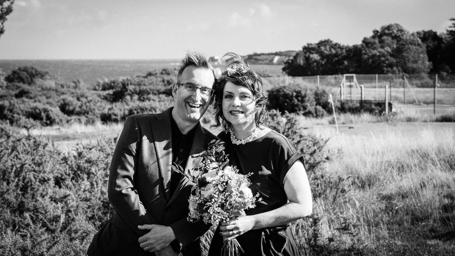 Black and white photo of a smiling couple outdoors, woman holding a bouquet, with scenic coastal landscape behind them.