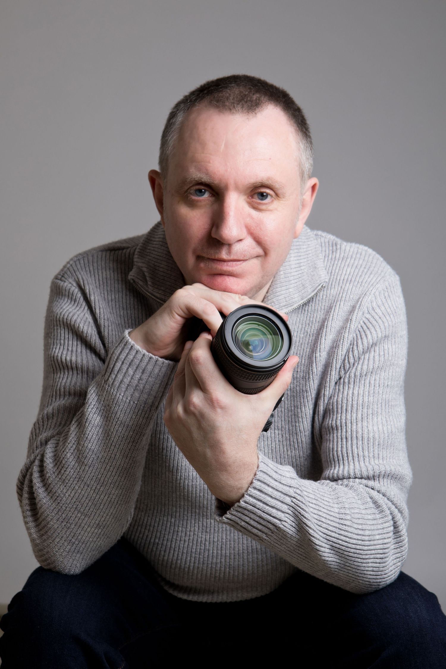 Male photographer holding camera lens, smiling in grey sweater, studio portrait shot.