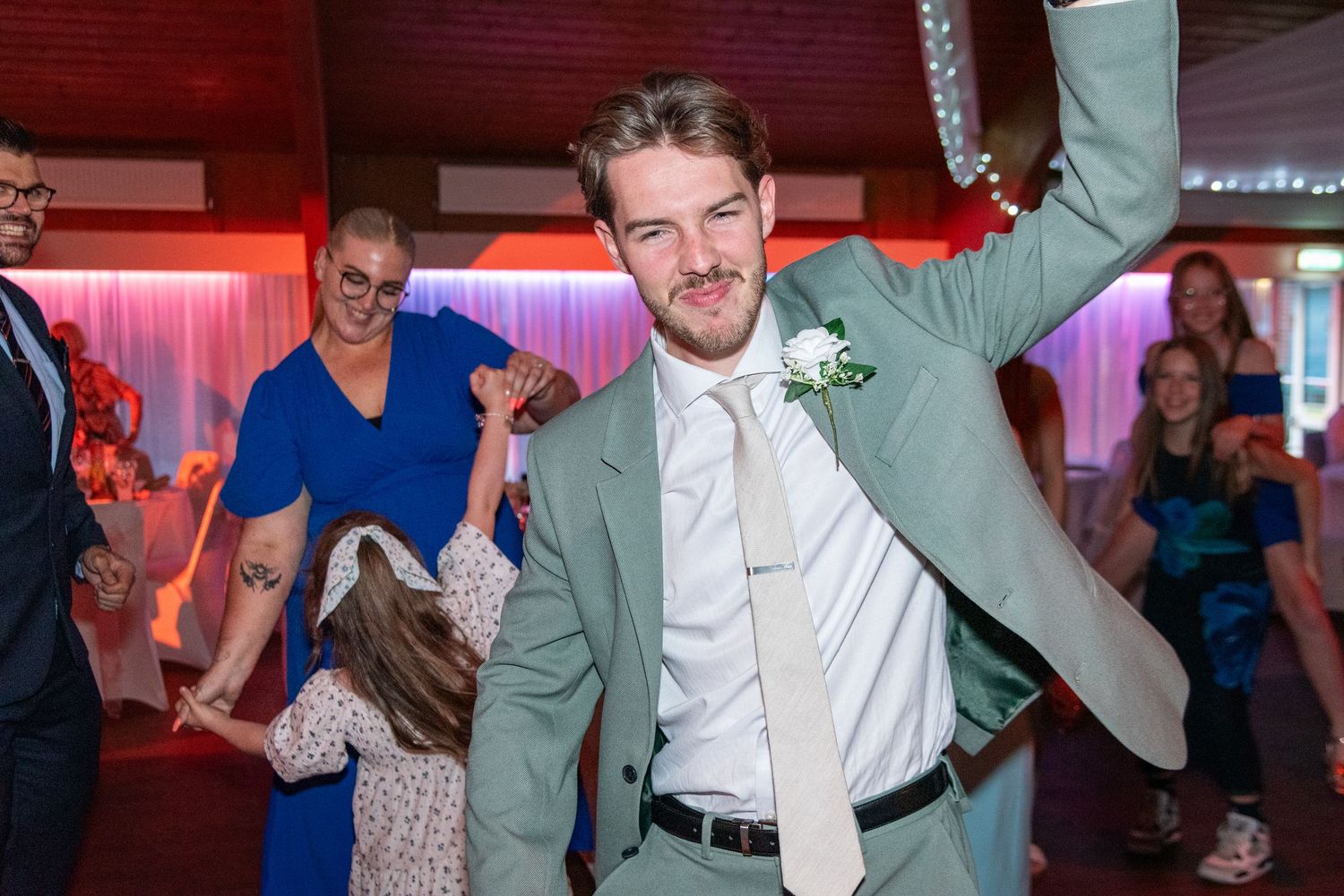 Groom in mint green suit dancing energetically at wedding reception with guests celebrating in background.