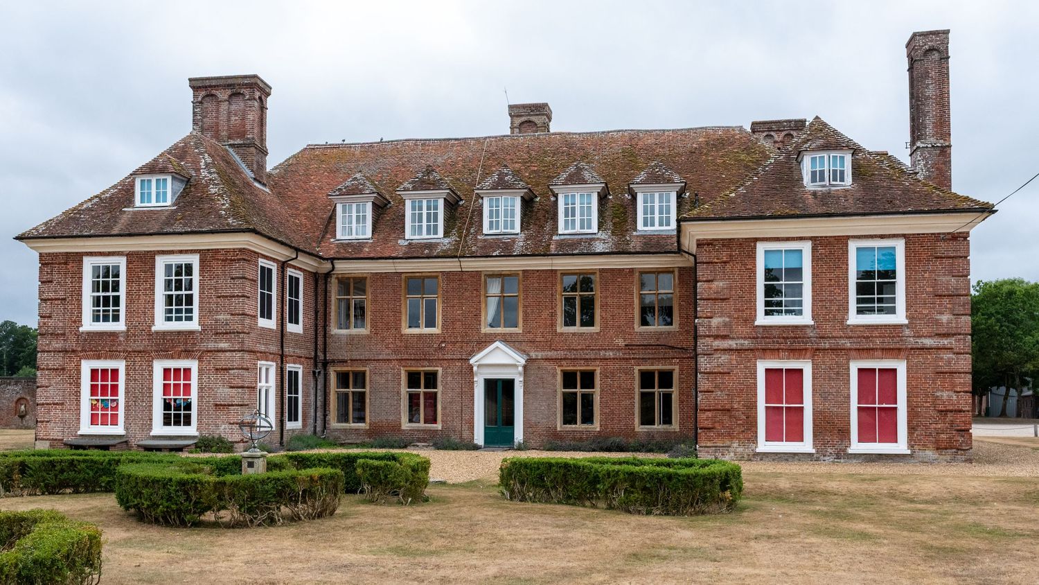 Historic red brick Georgian manor house with white-framed windows, red doors, manicured hedges, and a grand symmetrical facade.