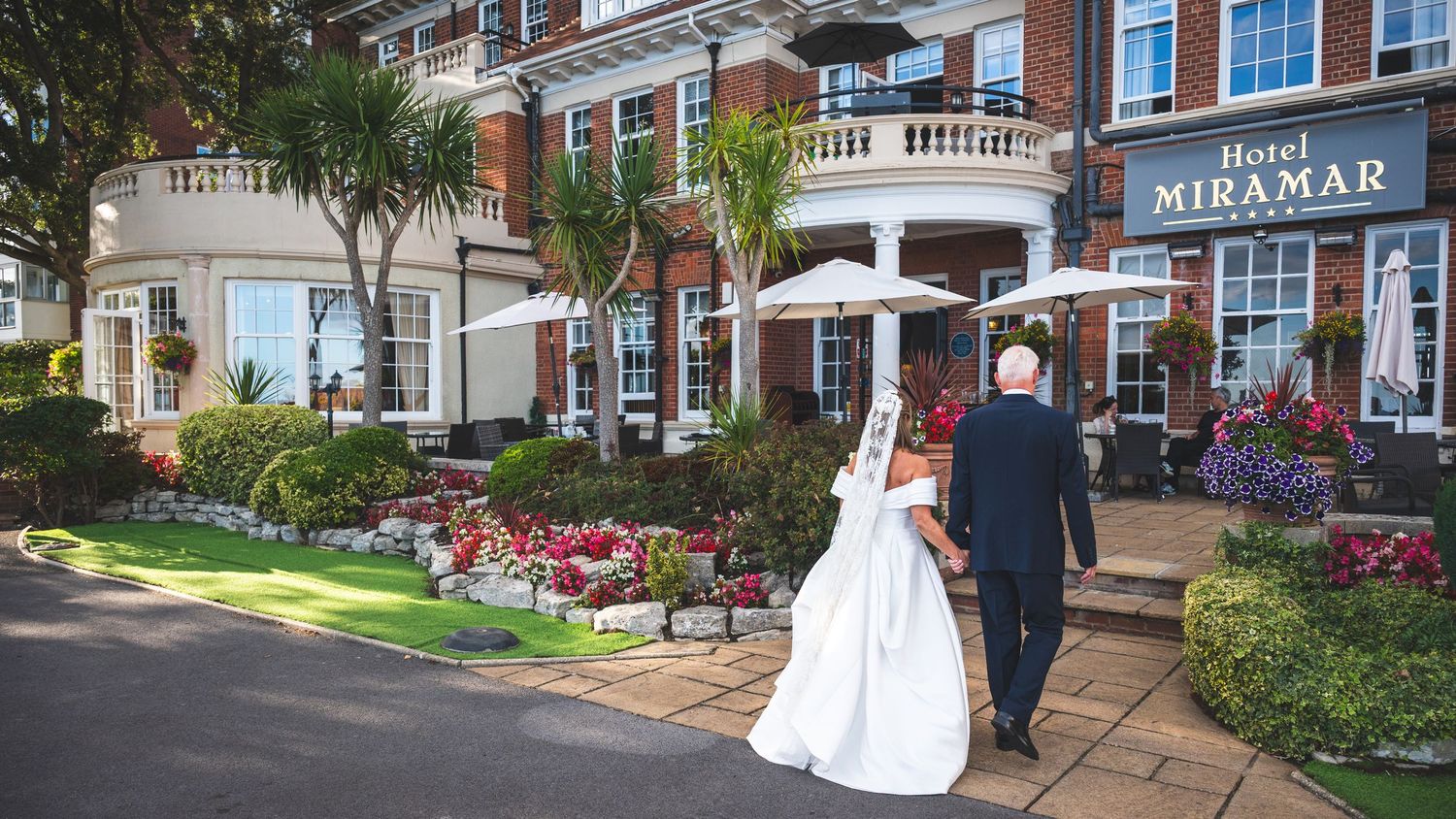 Bride in a long white gown walking hand in hand with a groom in a dark suit toward the Hotel Miramar, with palm trees and flower beds.