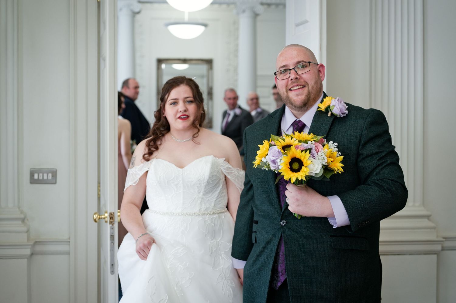 Bride in white gown and groom in green suit holding sunflower bouquet walk together in elegant hall.