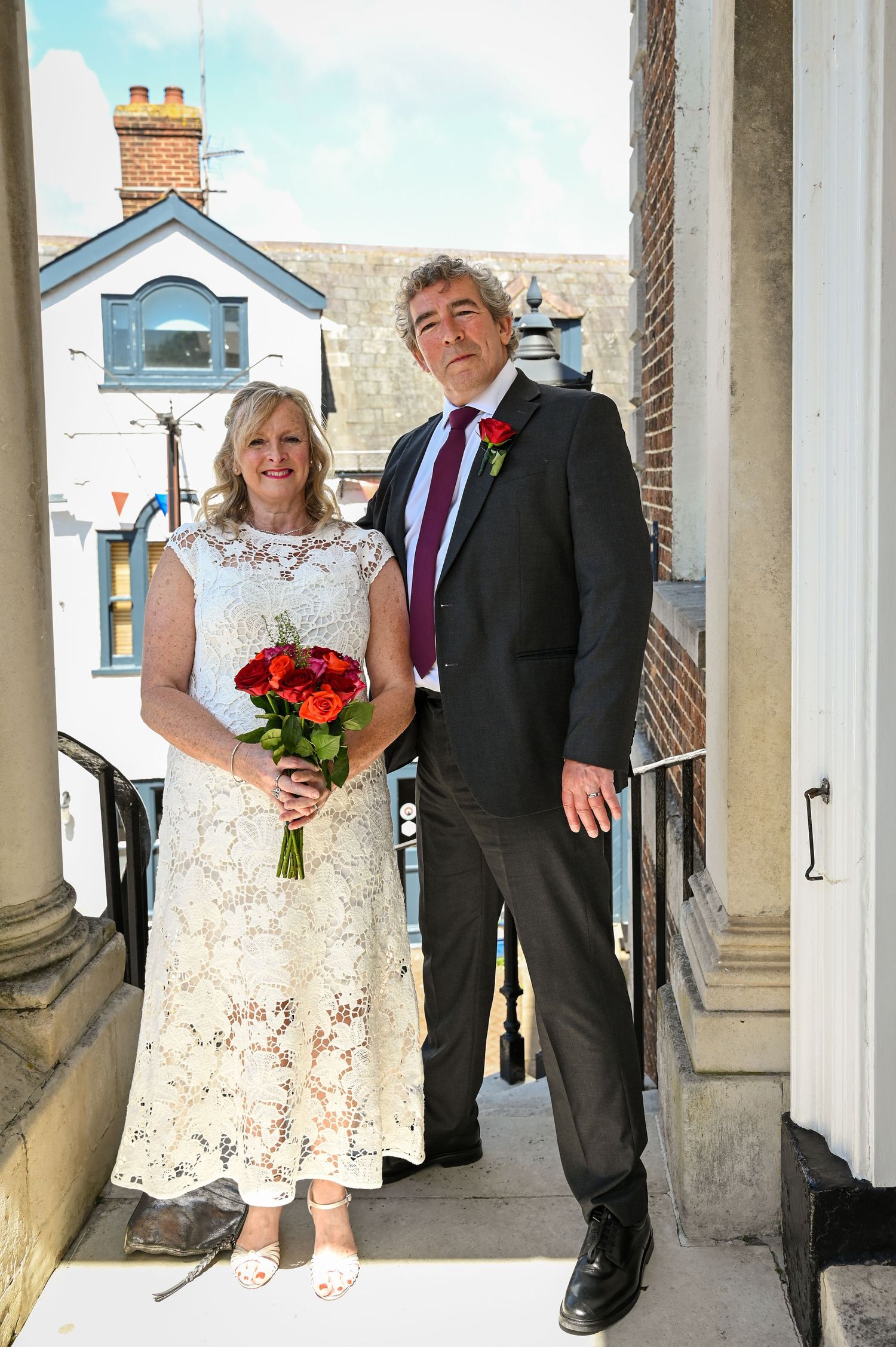 Smiling bride in white lace dress holding red roses bouquet stands with groom in dark suit at outdoor wedding venue.