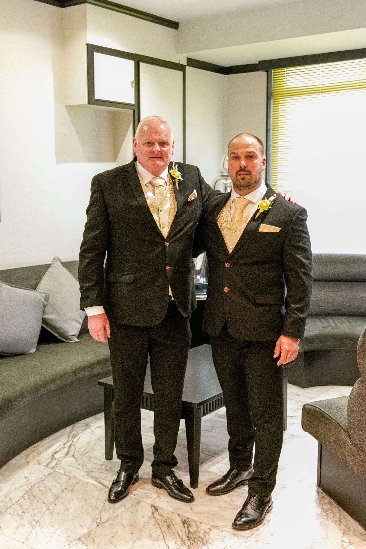 Two men in matching dark suits with gold ties and boutonnieres posing together in an elegant hotel room.