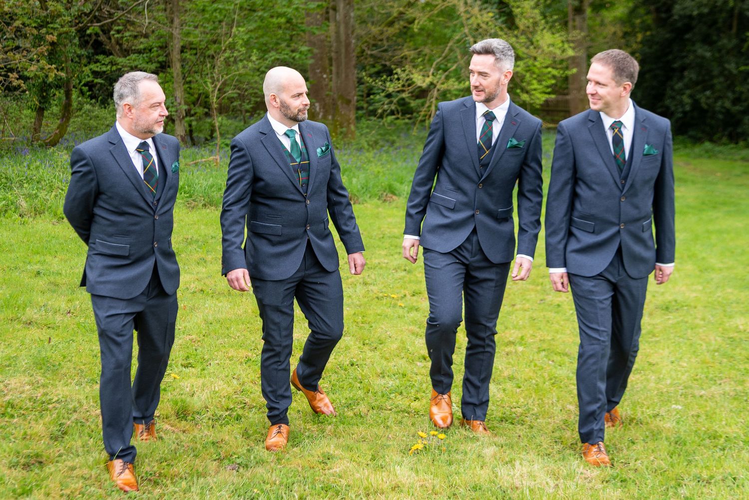 Four groomsmen in navy suits with brown shoes walking outdoors on green grass at a wedding.