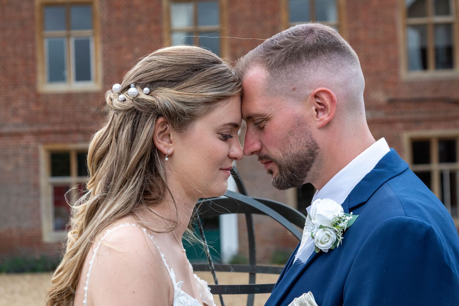 Bride and groom share an intimate forehead touch moment outdoors near a brick building on their wedding day.