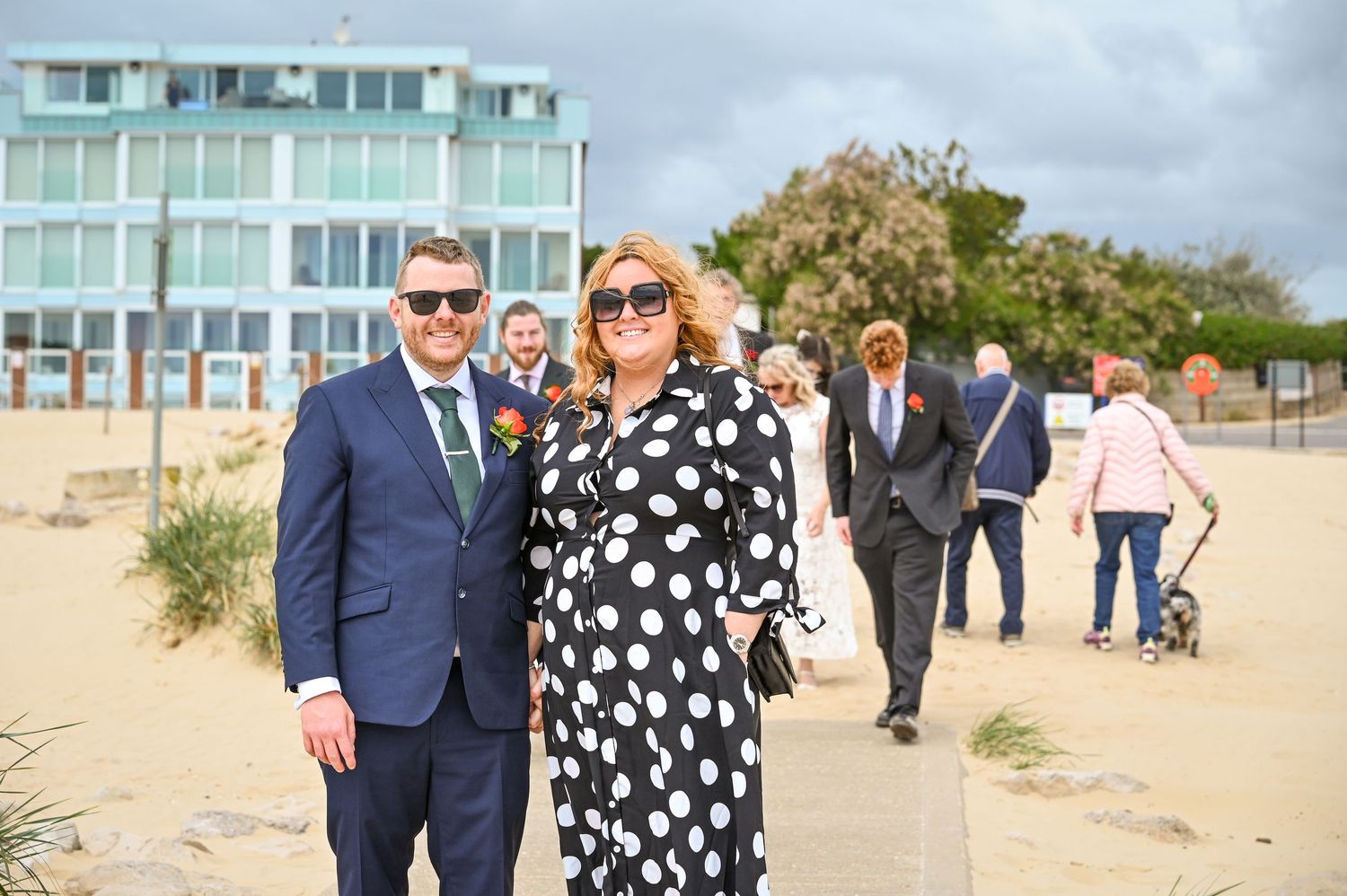 Couple in formal attire posing on a beachside path, woman in polka dot dress, man in navy suit, guests walking behind.