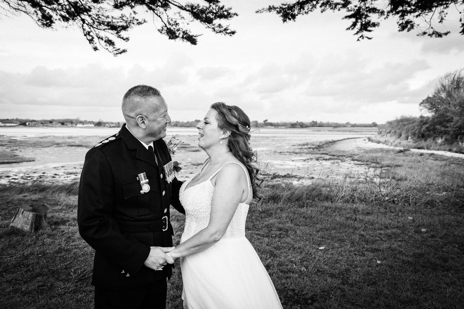 Black and white photo of a couple in wedding attire holding hands and smiling at each other near a scenic waterfront.