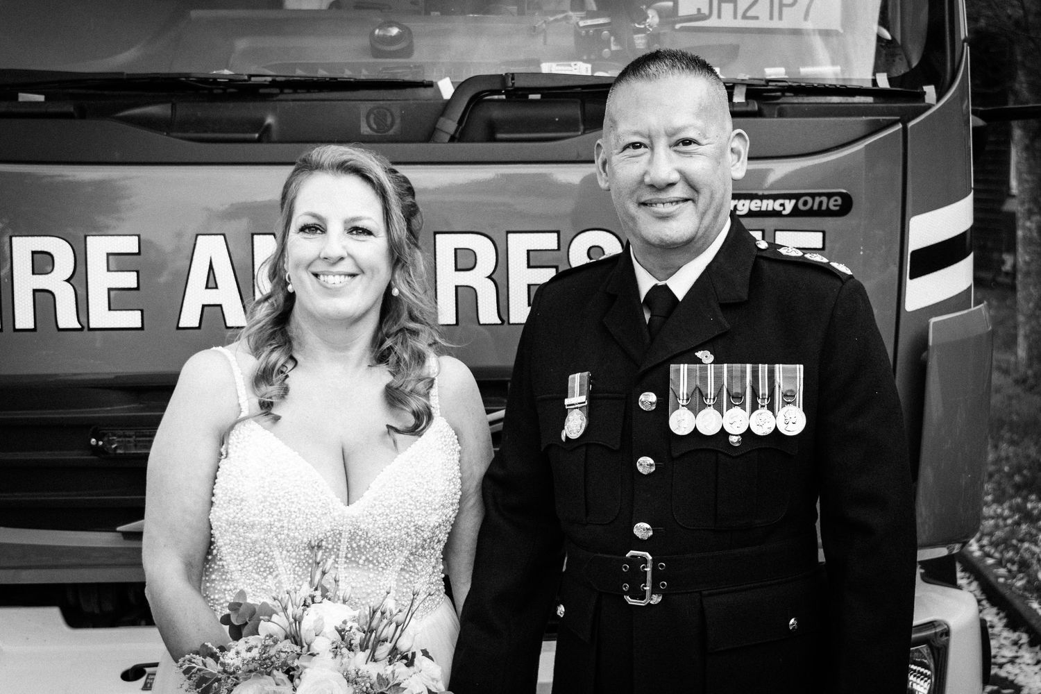 Black and white wedding photo of a bride and uniformed officer with medals posing in front of a fire rescue vehicle.