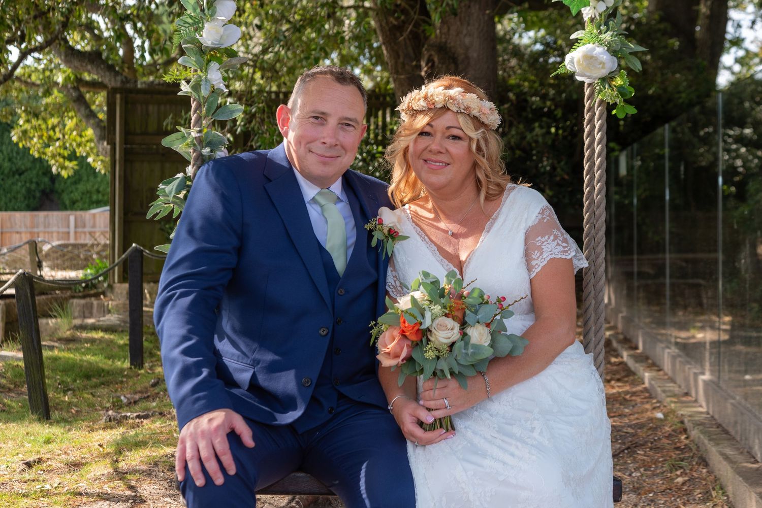 A couple poses in wedding attire outdoors, groom in blue suit, bride in white lace dress holding a floral bouquet.