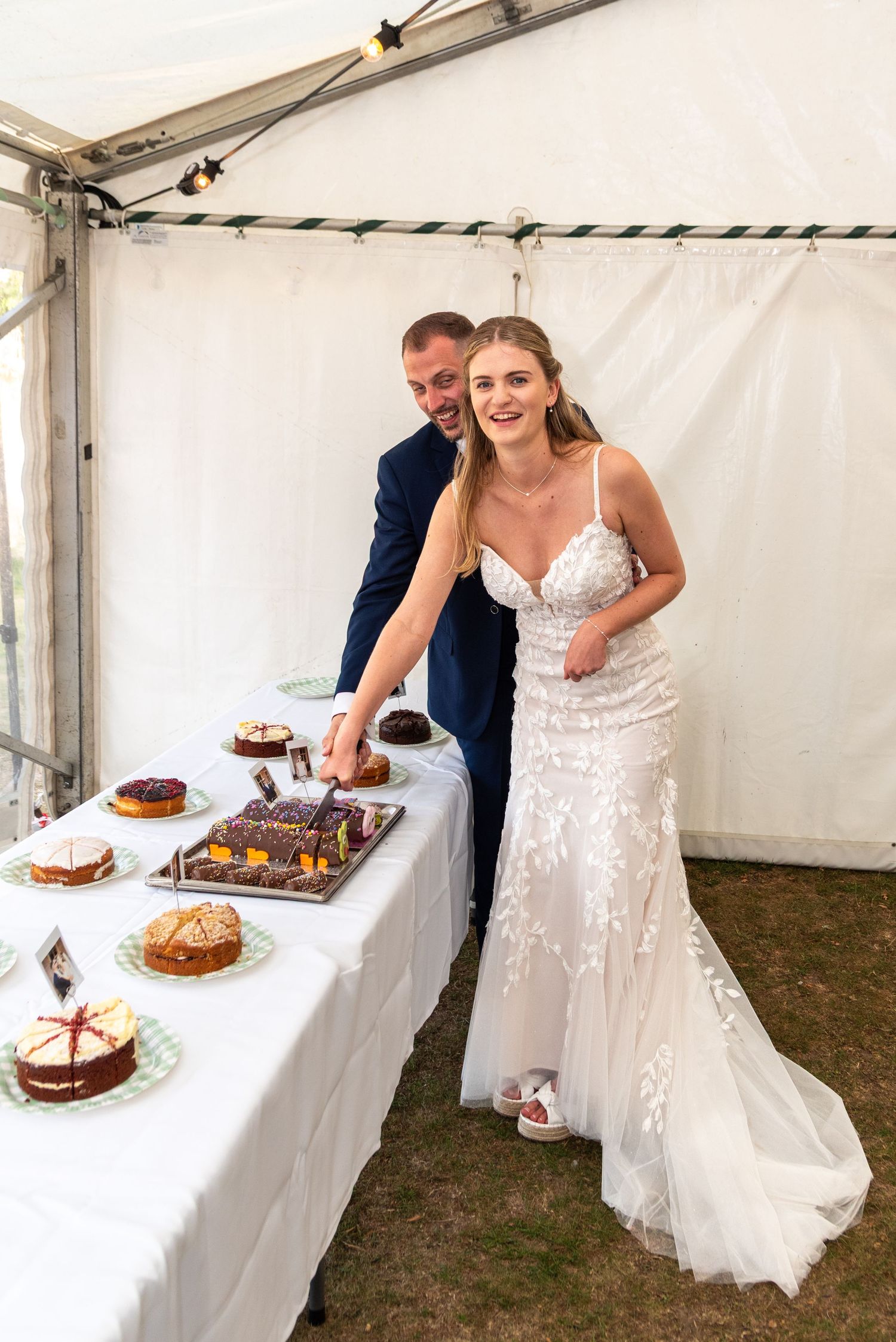 Smiling bride and groom in navy suit cut wedding cake at table inside marquee tent.