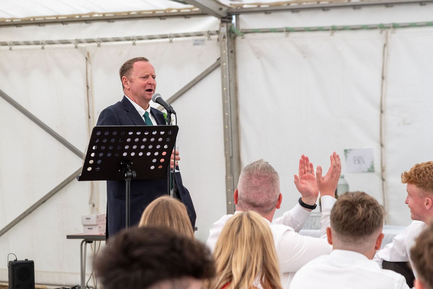Man in dark suit speaking at podium during wedding reception in white marquee tent.